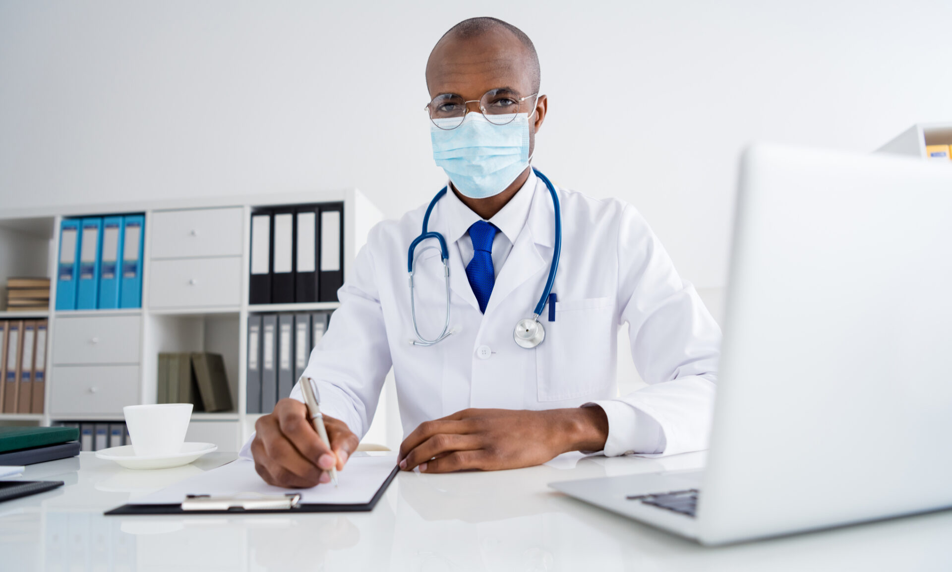 Masked doctor writing on clipboard at desk